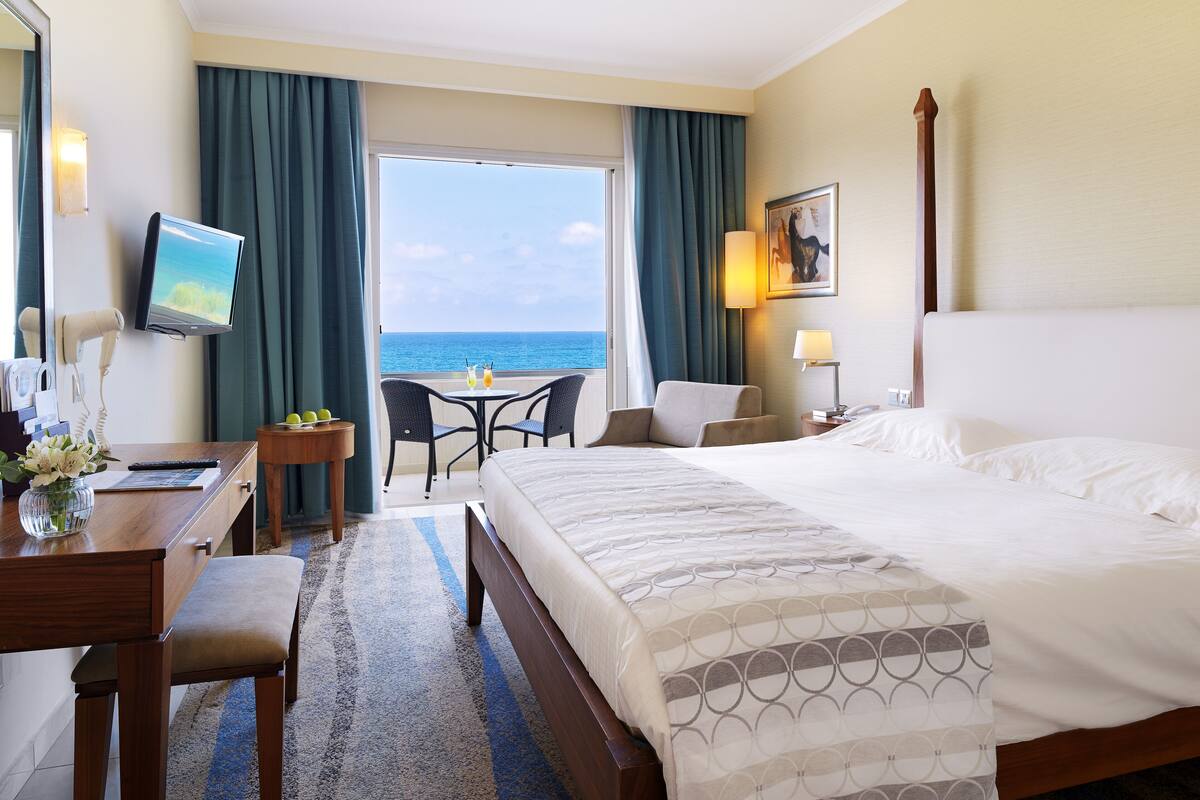 A hotel room with a bed, a desk with a chair, a TV, a table with two chairs, and a view of the sea.