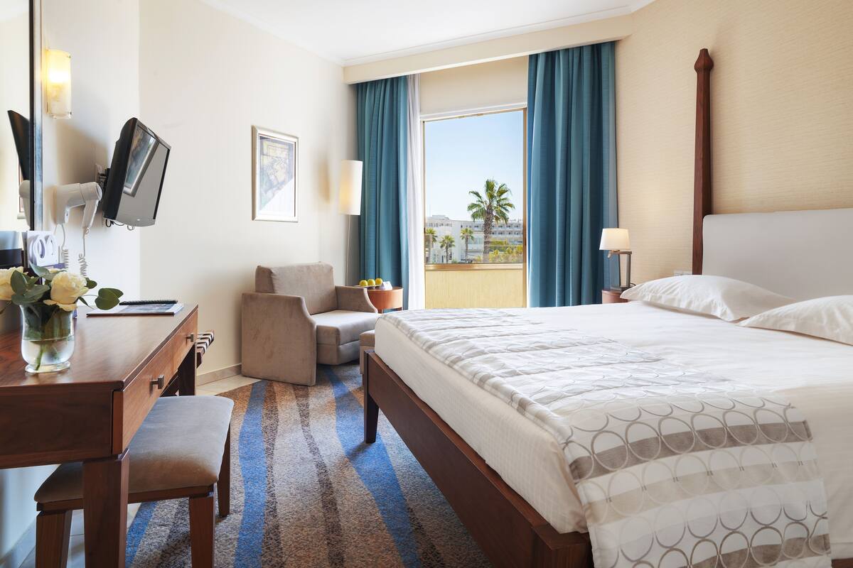 A hotel room with a bed, desk, chair, TV, and a view of palm trees.