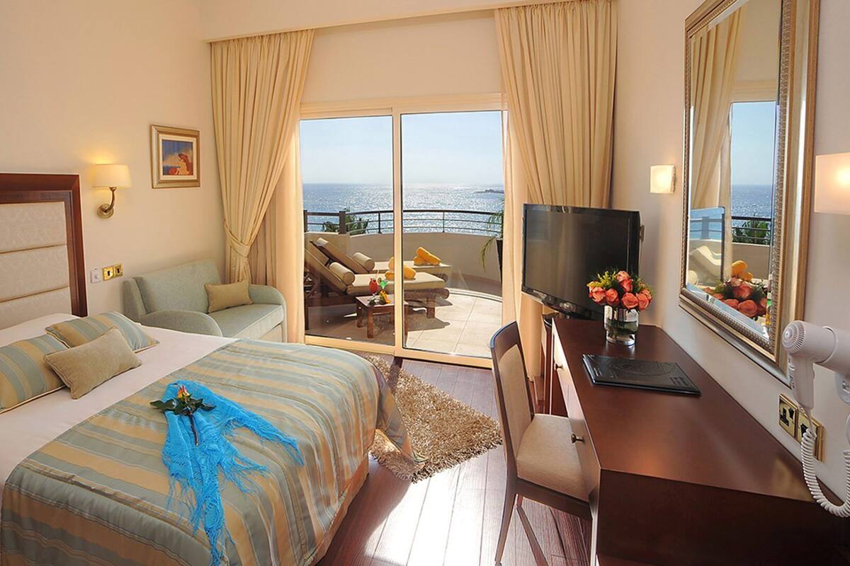 A hotel room with a bed, a desk with a TV, a chair, and a view of the sea through the balcony doors.