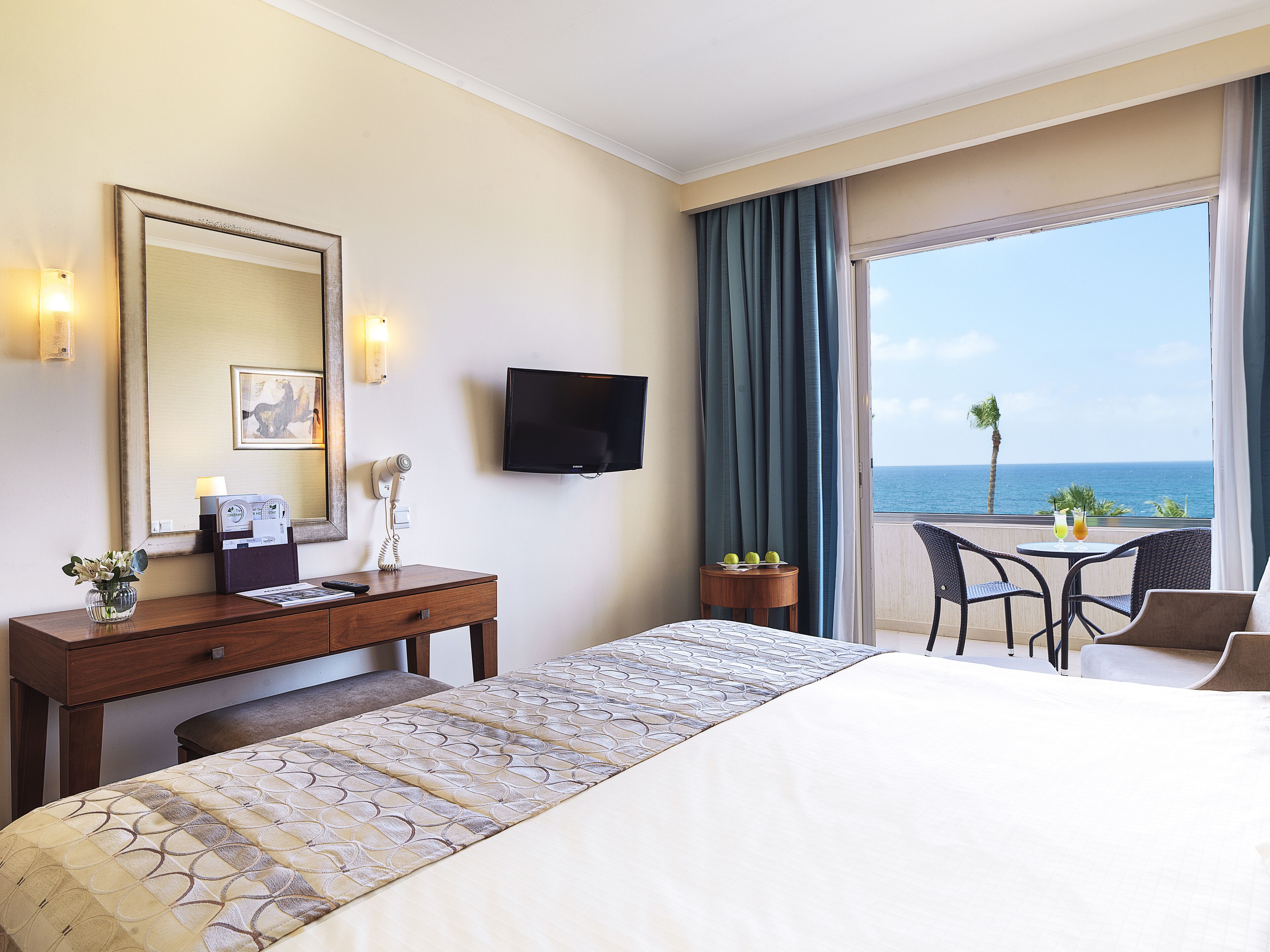 A hotel room with a bed, a desk with a hairdryer, a TV, and a balcony with a view of the sea.