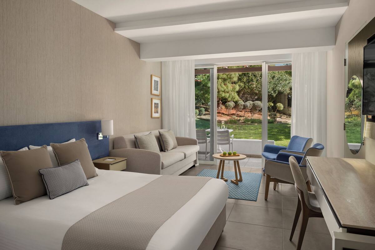 Habitación de hotel moderna con una cama grande, un sofá, una mesa de comedor y vistas a un jardín ajardinado a través de la puerta de vidrio.