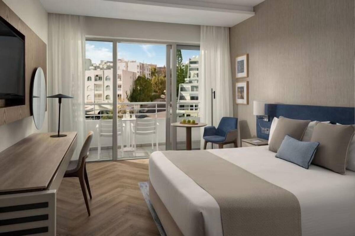 Habitación de hotel con cama, escritorio, silla, mesita pequeña y balcón con vista a edificios.