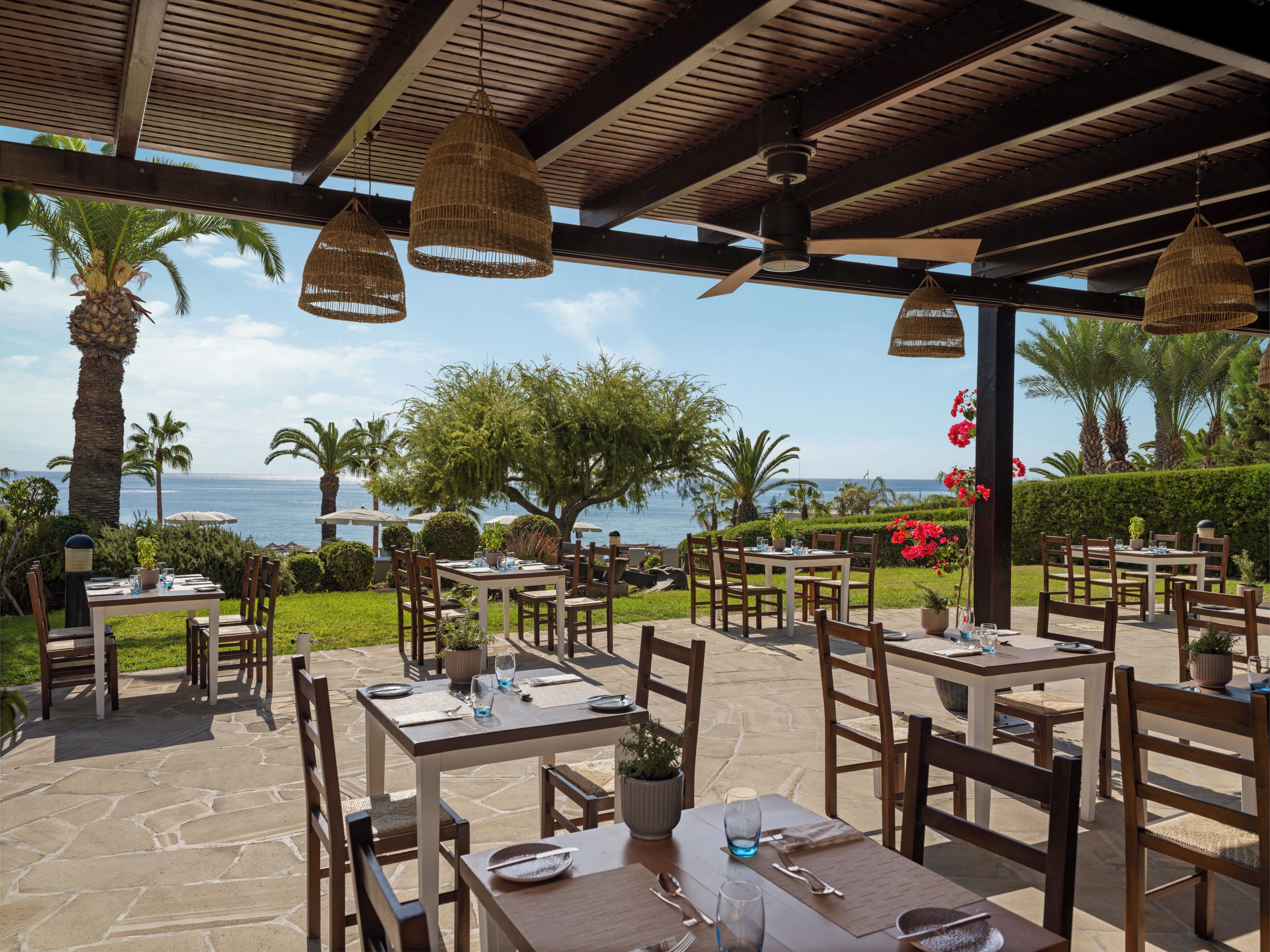 Un área de comedor al aire libre con mesas y sillas, una pérgola y vistas al mar.