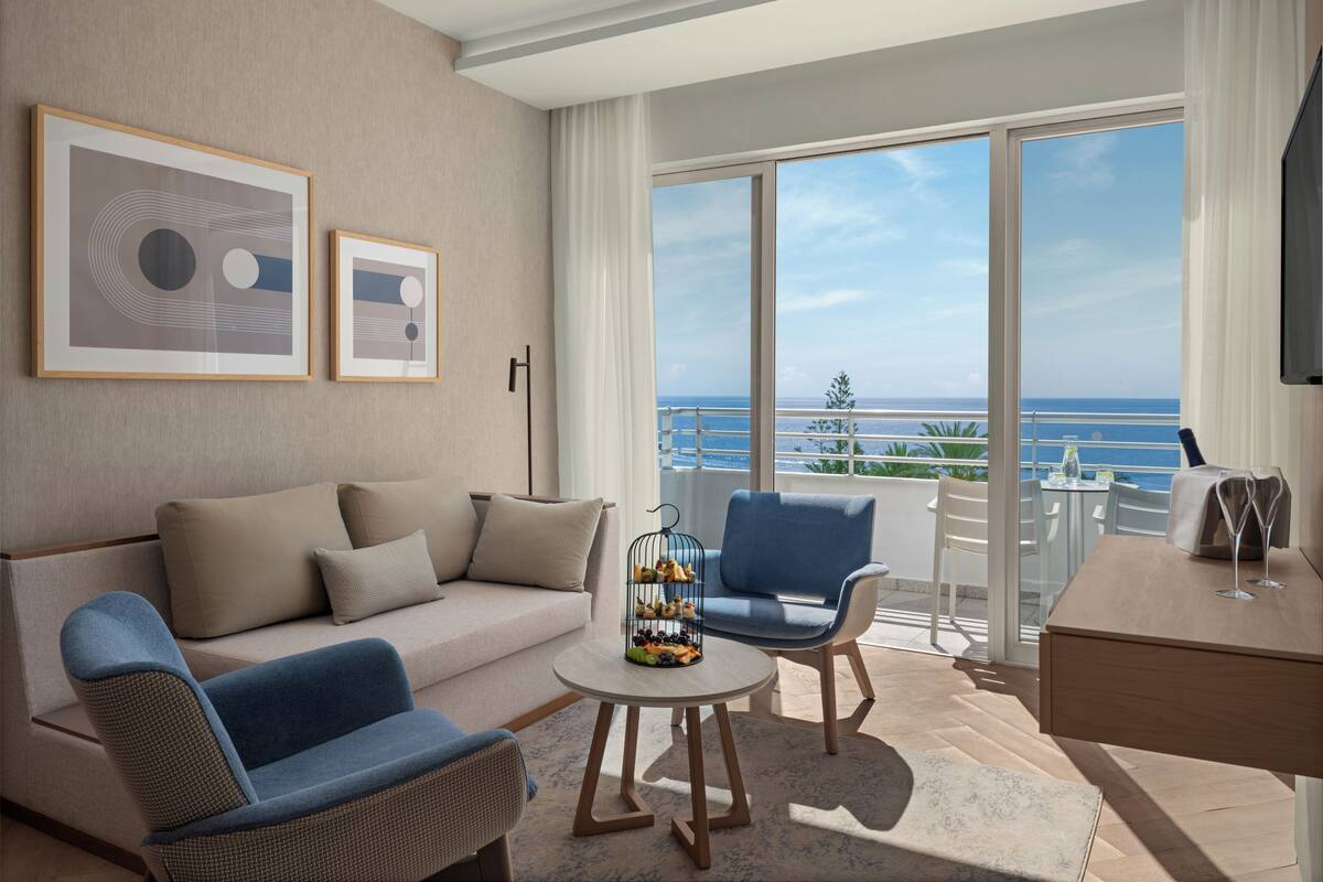 Una habitación de hotel moderna con balcón, un sofá, una silla, una pequeña mesa con fruta y obras de arte en la pared.