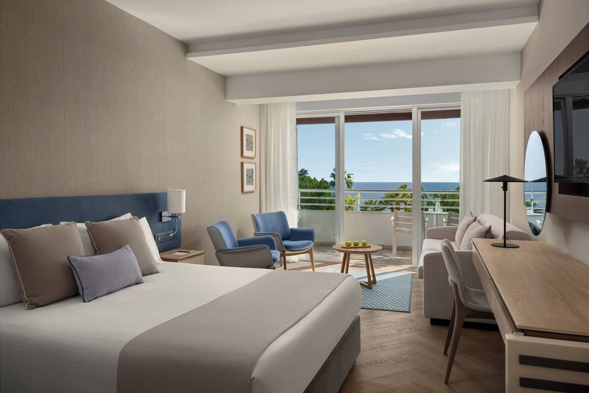 Habitación de hotel moderna con una cama grande, un escritorio, una silla y un balcón con vista al océano.