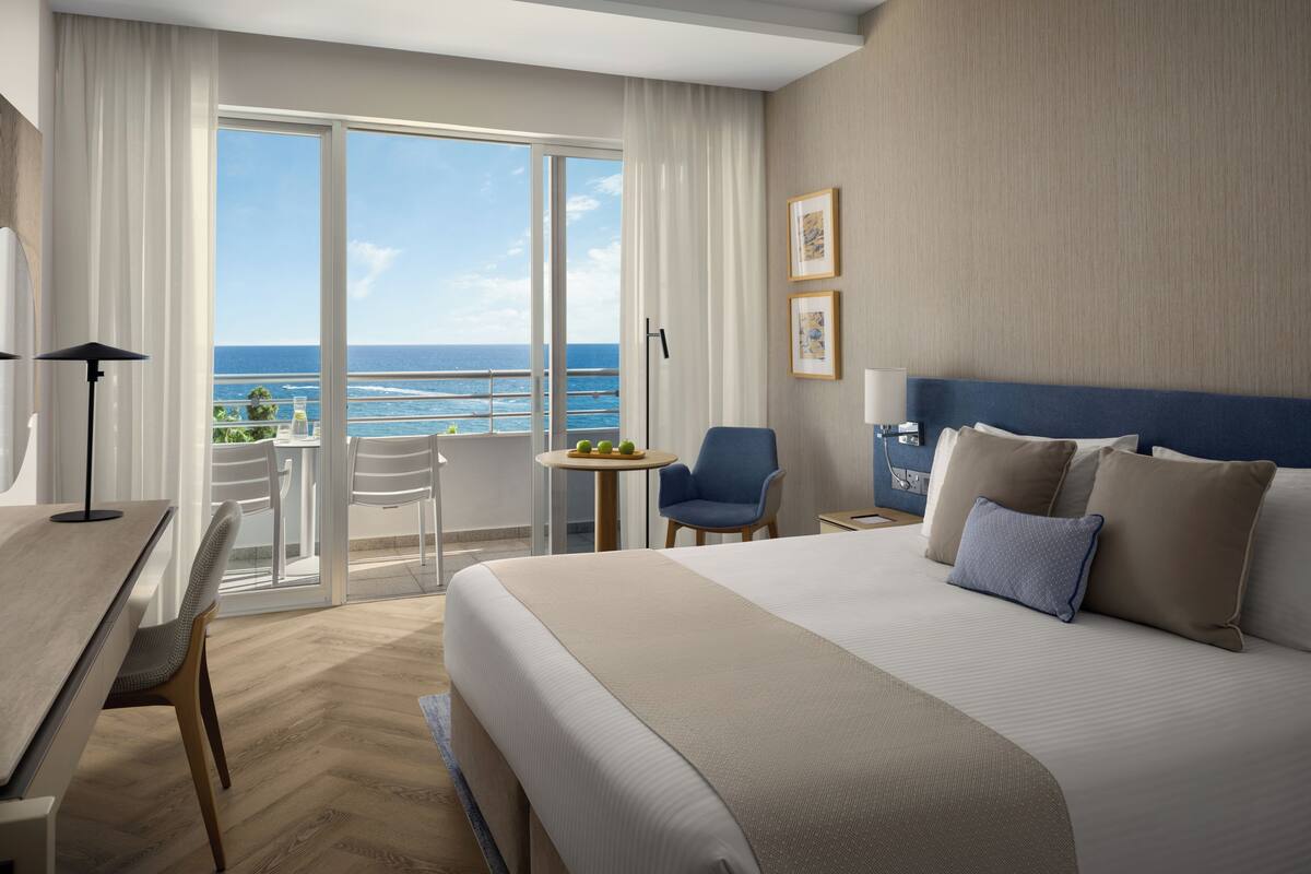 Habitación de hotel con cama, escritorio, silla, una pequeña mesa con fruta y vista al océano.