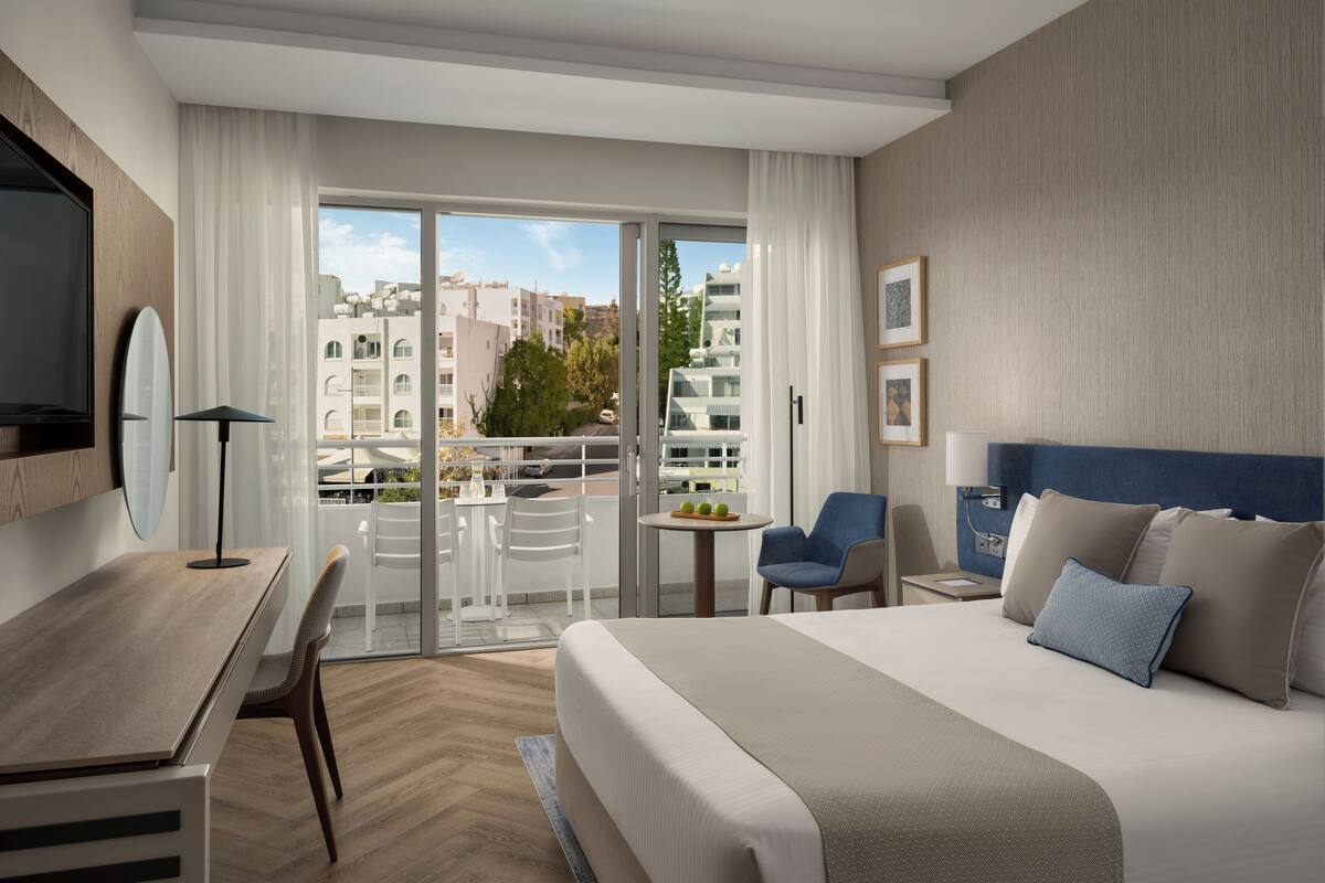 Habitación de hotel con cama, escritorio, silla, mesa y balcón con vista a edificios.