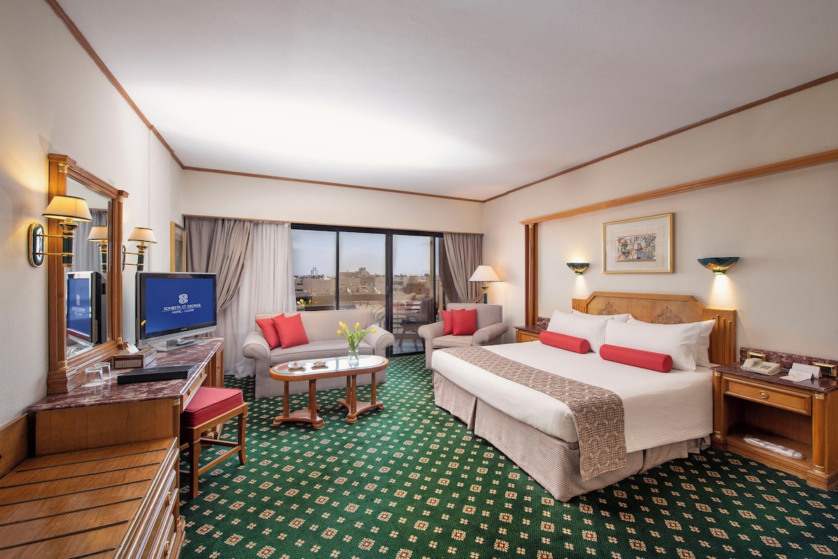 Quarto de hotel com cama, escrivaninha com computador, sofá, mesa de centro e vista da cidade.