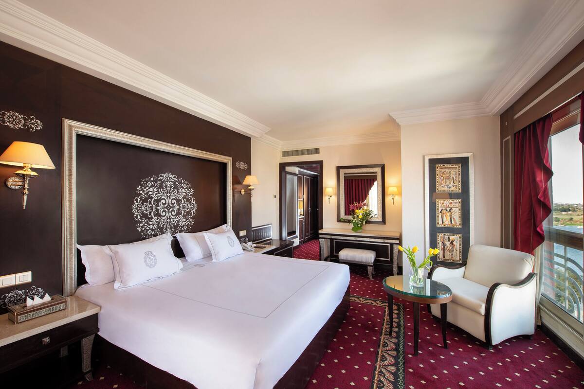 Quarto de hotel com uma cama grande, uma escrivaninha com cadeira, uma mesinha com um vaso e uma janela ampla com cortinas.