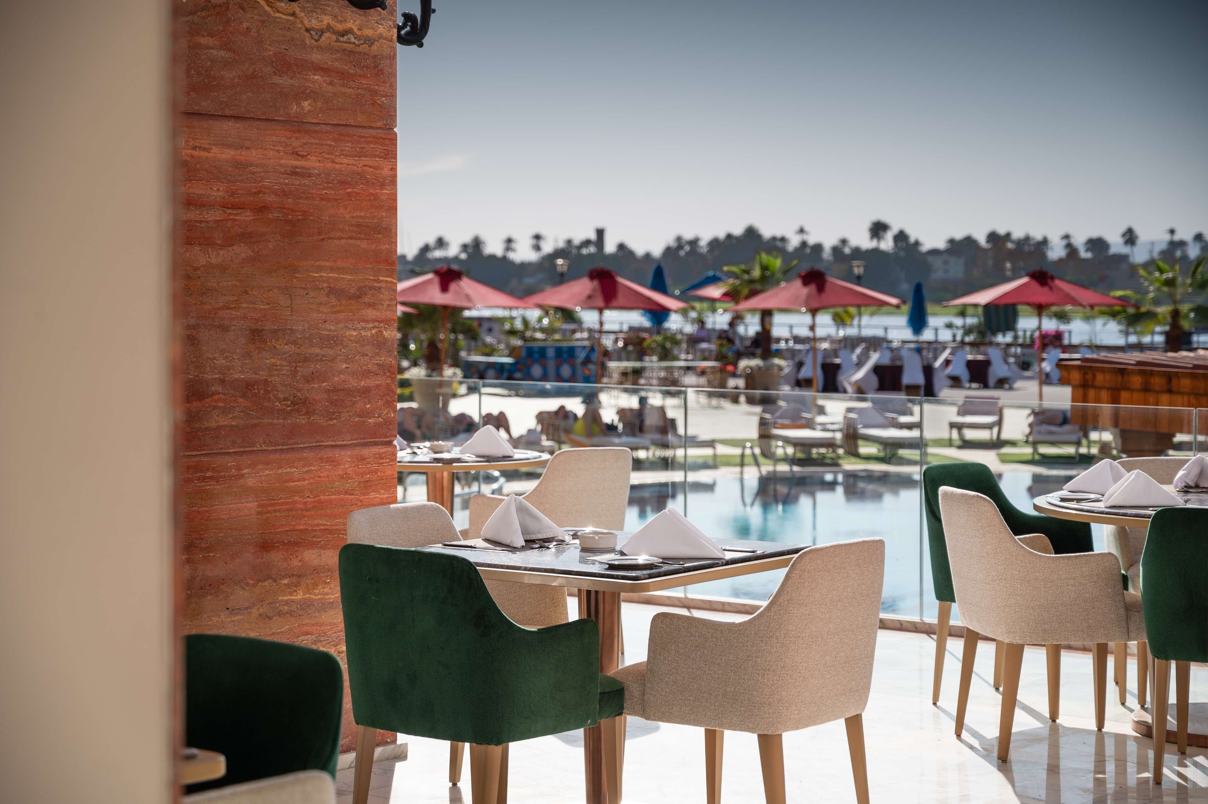 Un área de comedor junto a la piscina con mesas, sillas y sombrillas, y vistas a la piscina y palmeras.