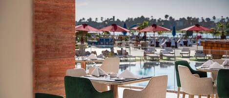 Un área de comedor junto a la piscina con mesas, sillas y sombrillas, y vistas a la piscina y palmeras.