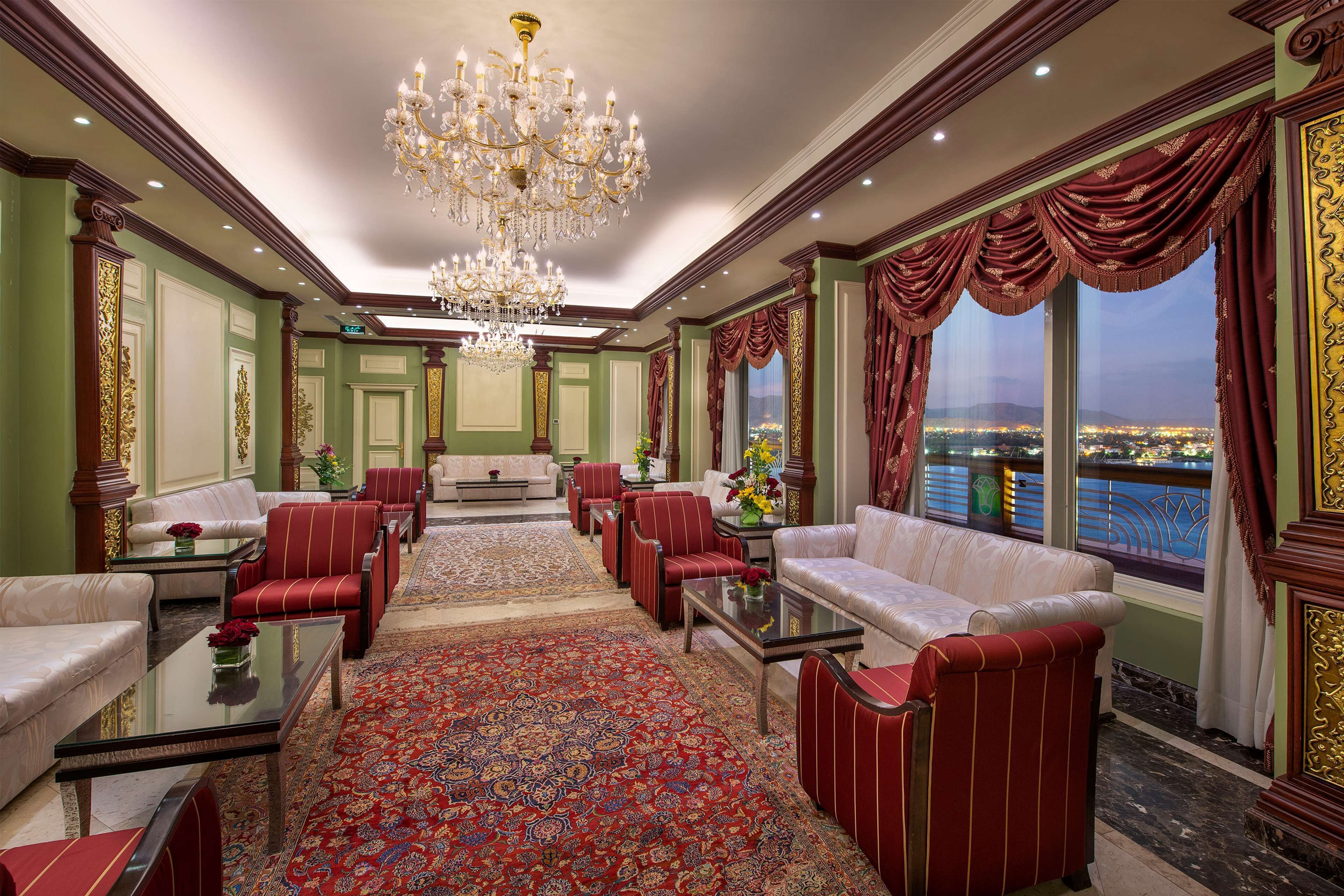 A luxurious lobby with a chandelier, red and white seating, a large rug, and a view of a cityscape.