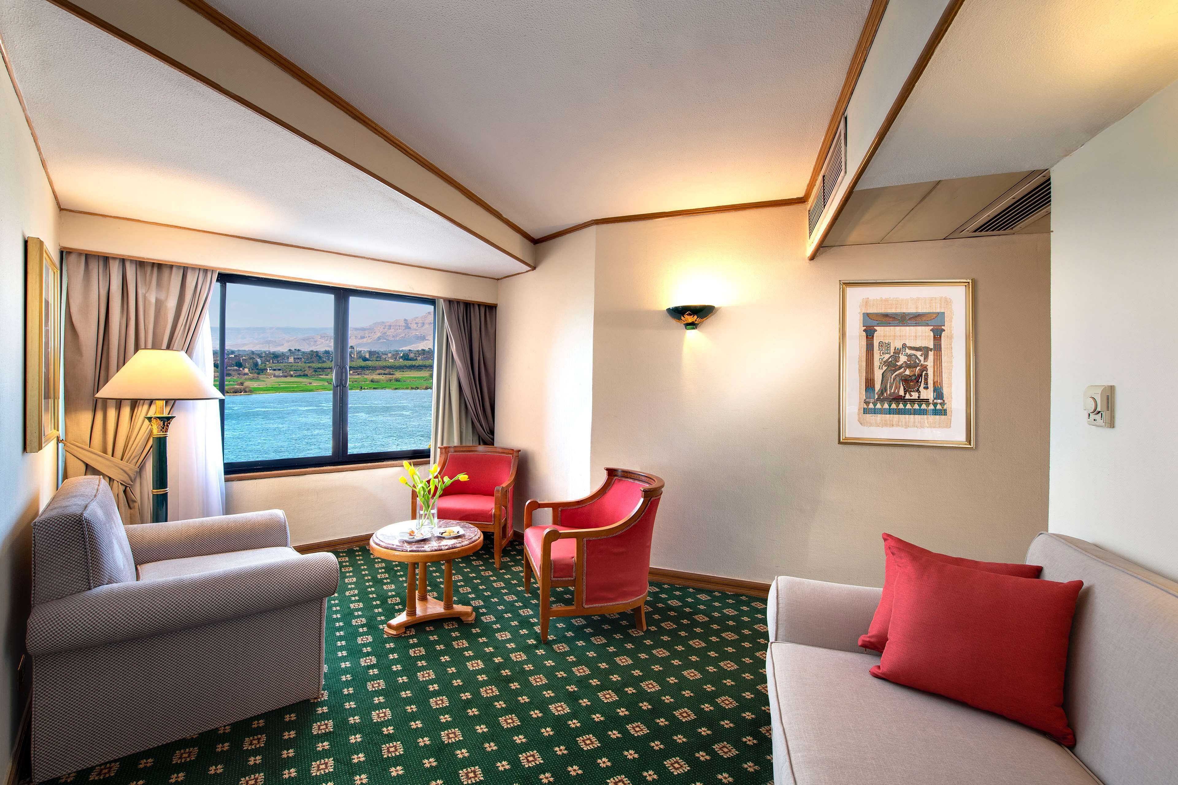 A hotel room with a large window, two red chairs, a grey sofa, and a green patterned carpet.