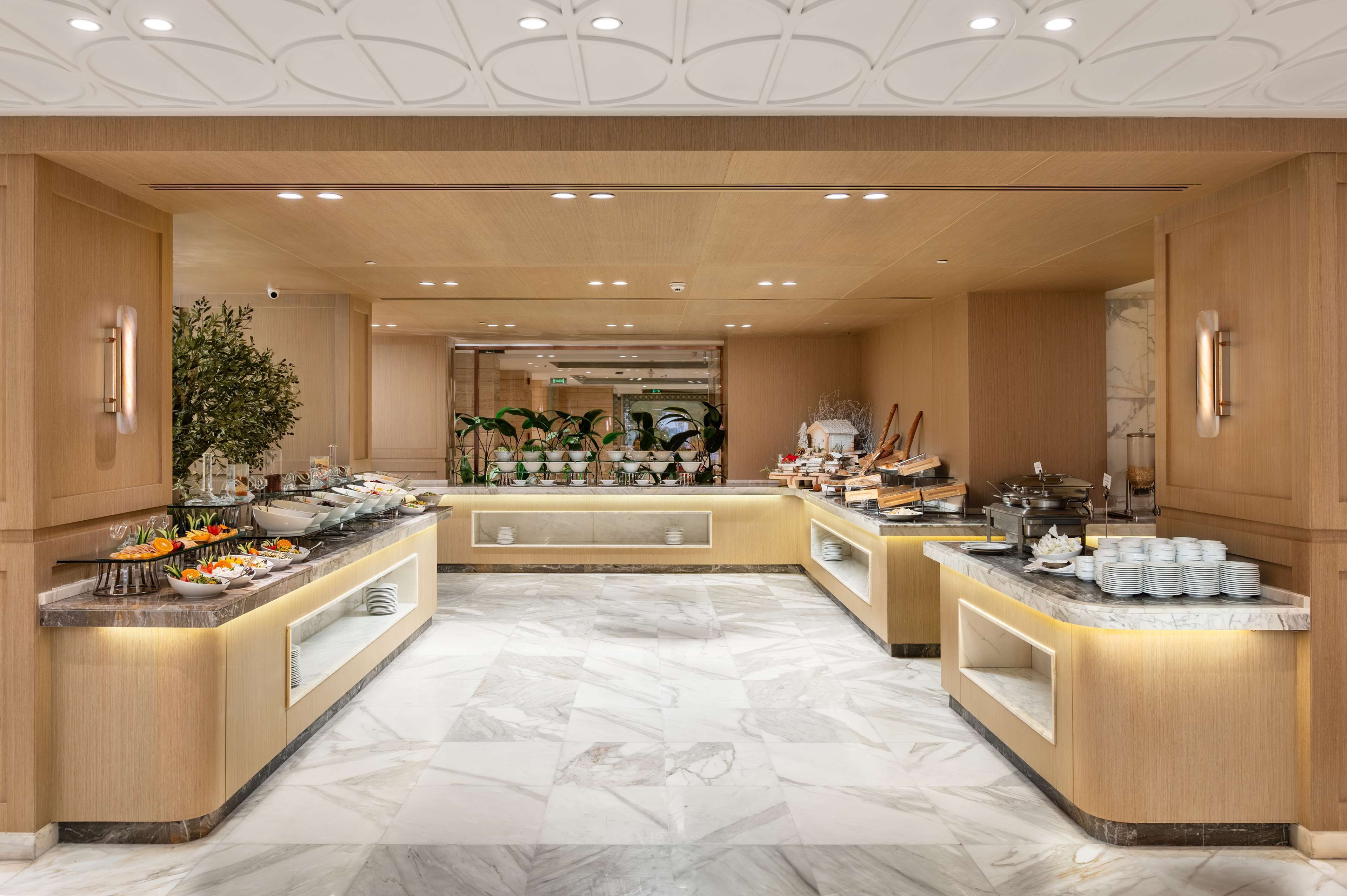 Un vestíbulo de hotel con suelo de mármol, mostrador de recepción de madera y una zona de buffet con diversas opciones de comida.
