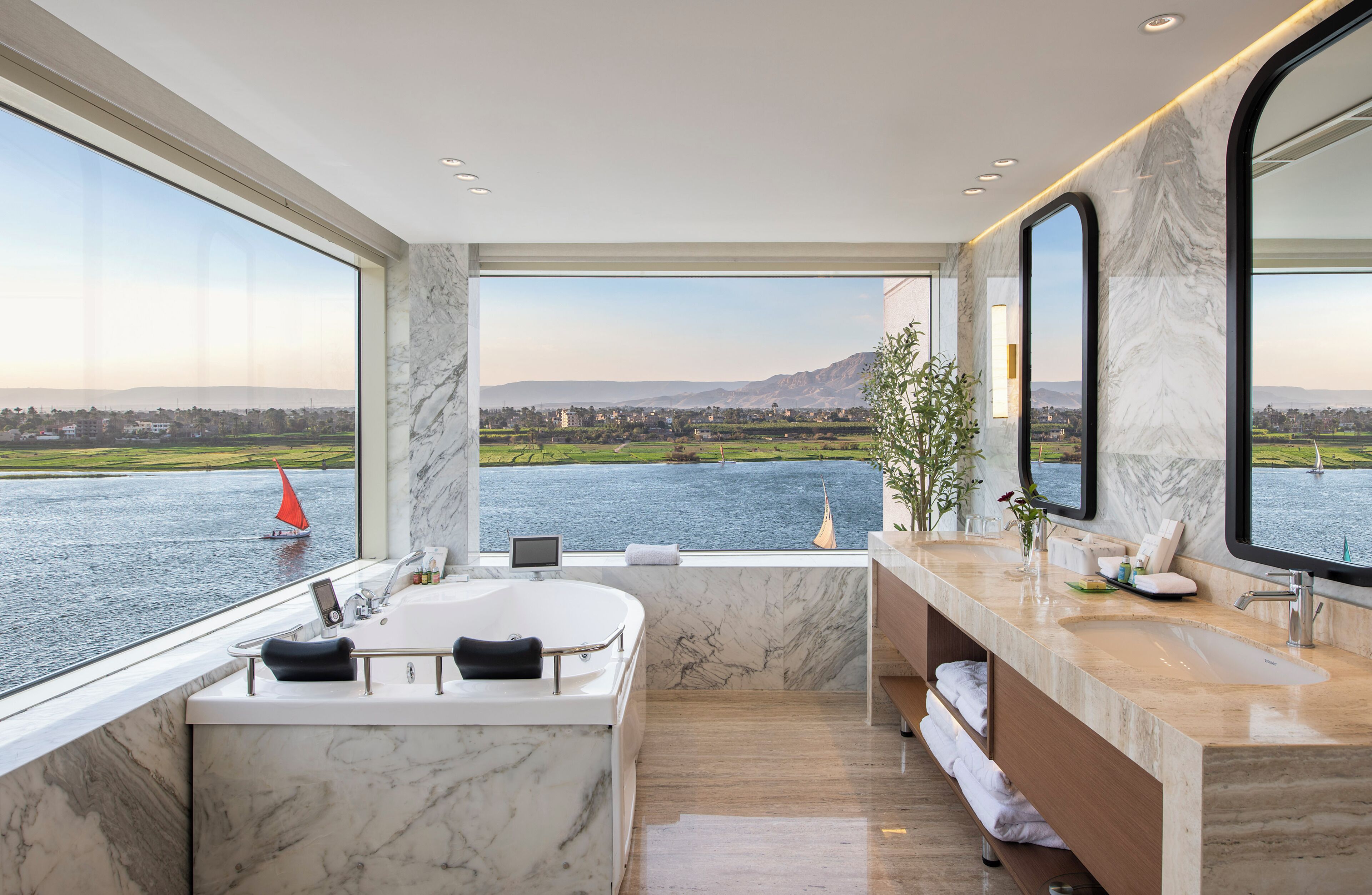 Un baño moderno con una bañera grande, un tocador con espejo y vistas a un lago con veleros.