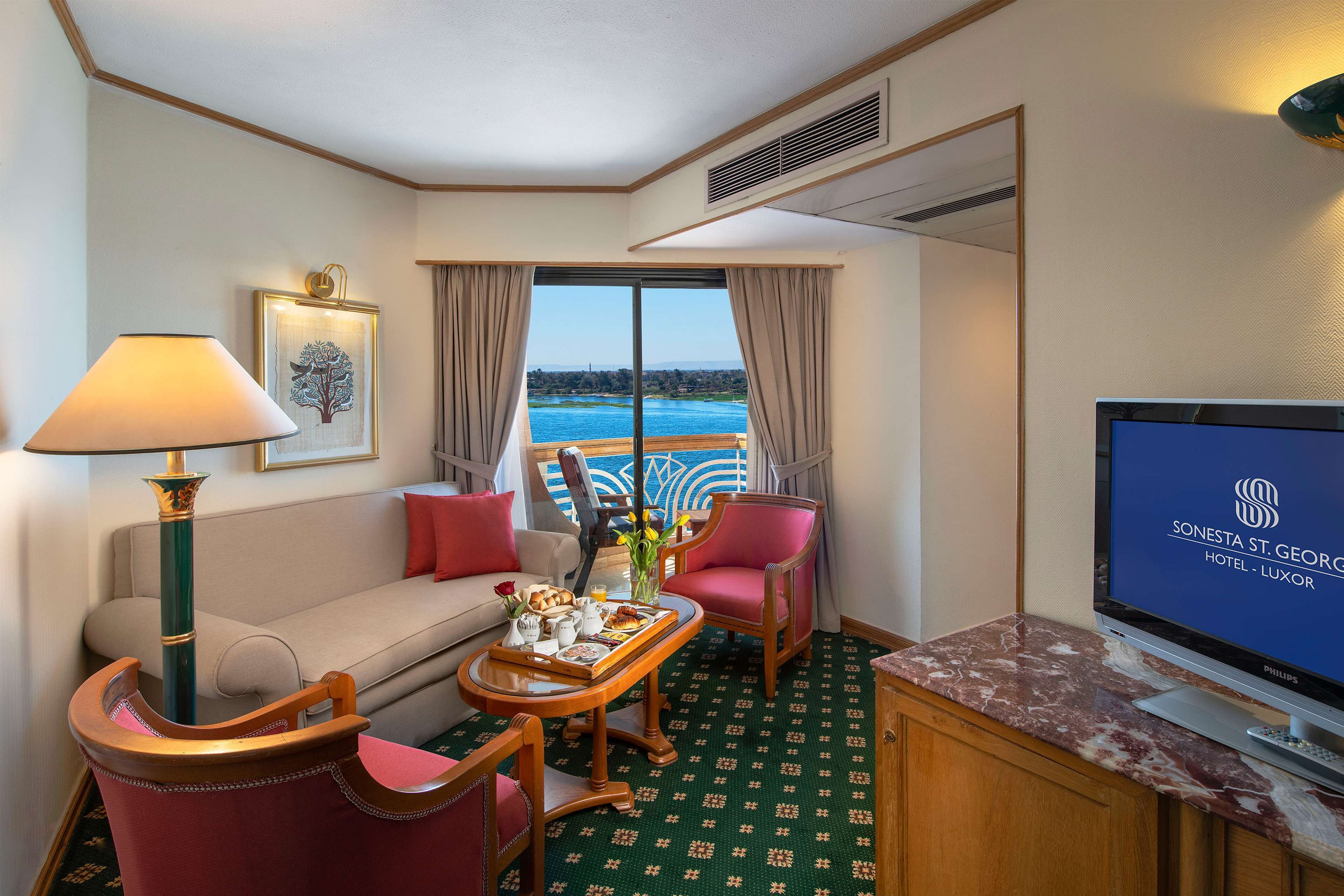 A hotel room with a sofa, two chairs, a coffee table with breakfast items, a TV, and a large window with a view of the sea.