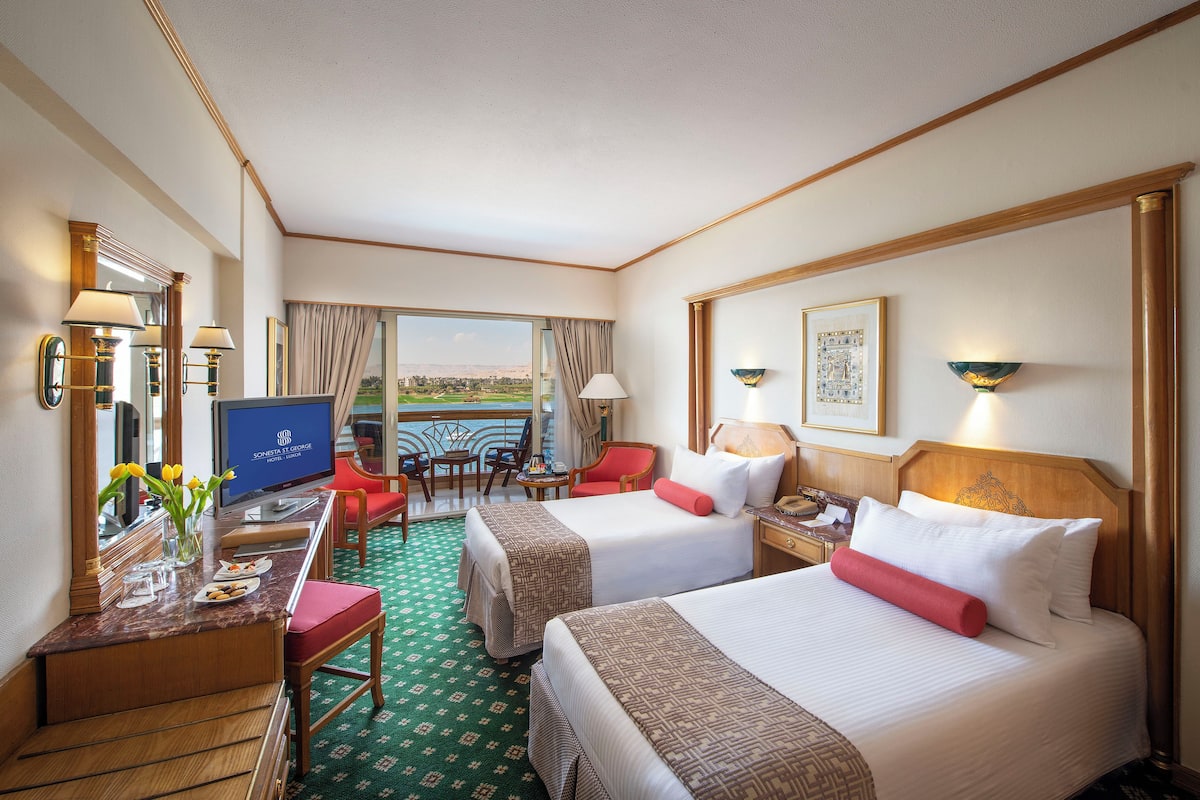 Quarto de hotel com duas camas, uma mesa com computador, uma cadeira, televisão e vista para o exterior.