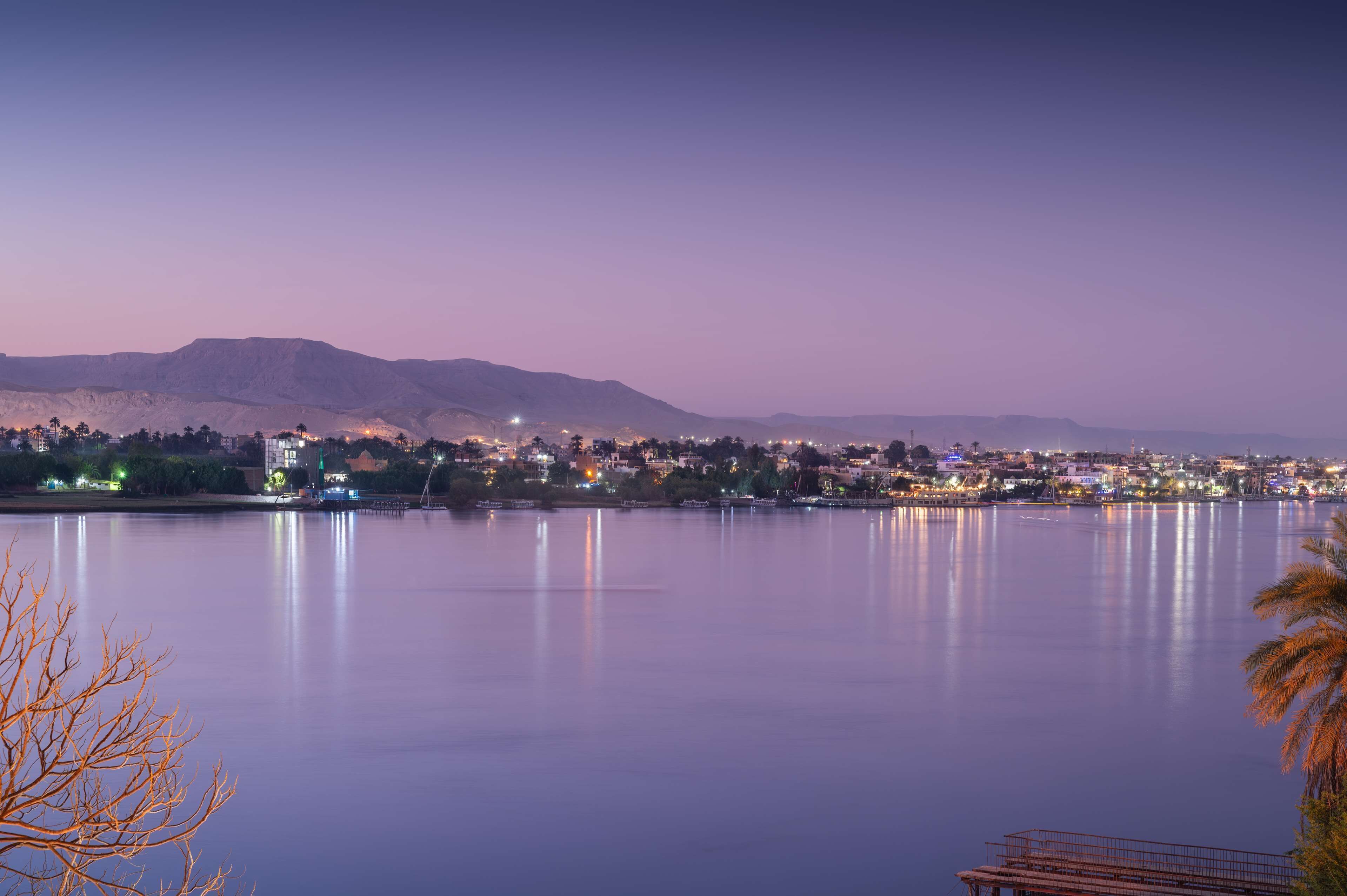 Un lago tranquilo con un horizonte urbano al fondo, destacando edificios iluminados y una cadena montañosa.
