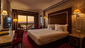 Habitación de hotel con una cama grande, un escritorio con televisor, un comedor y un balcón con vistas.