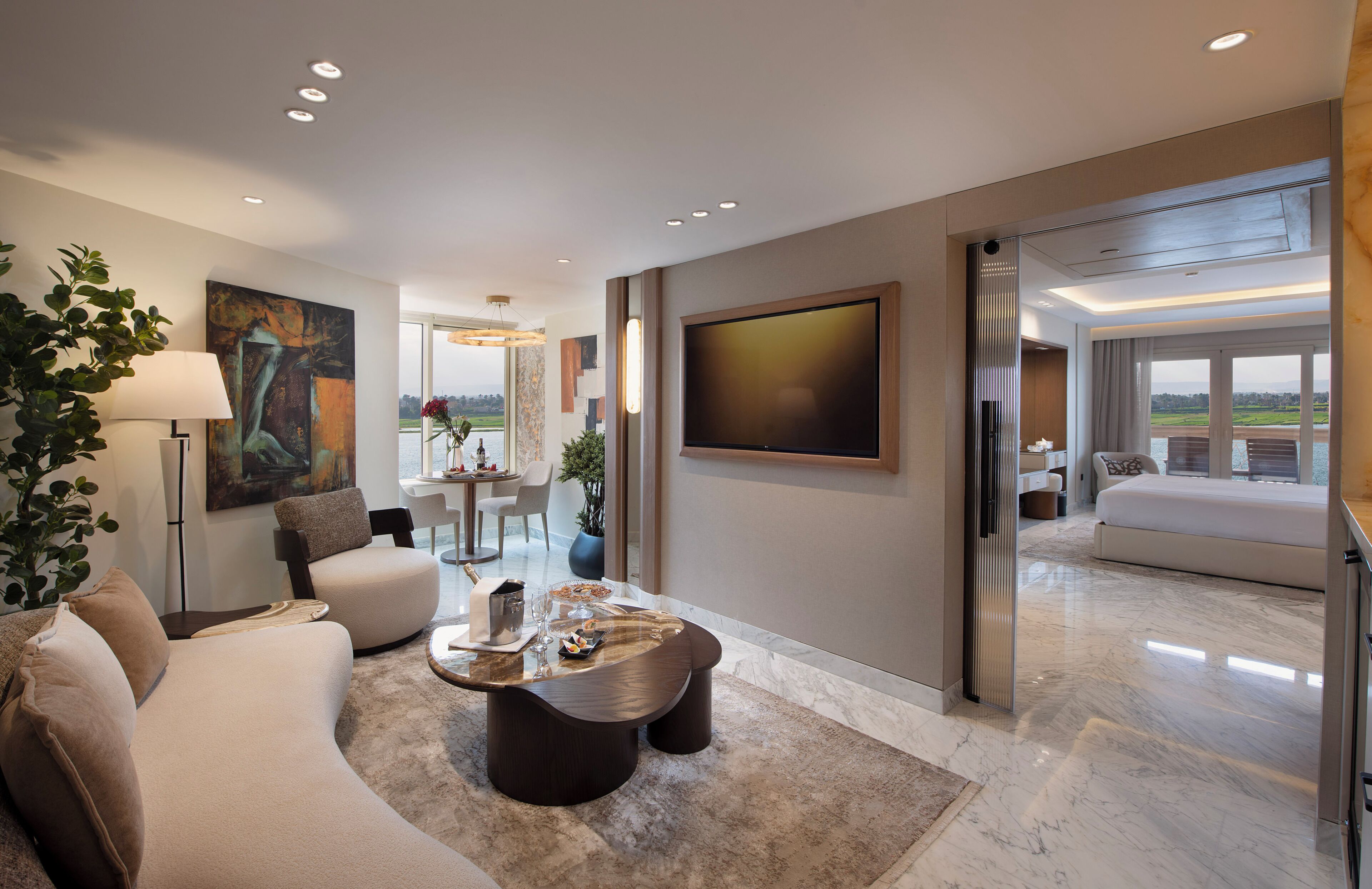 A modern hotel room with a large bed, a seating area with a coffee table, and a dining area with a view.
