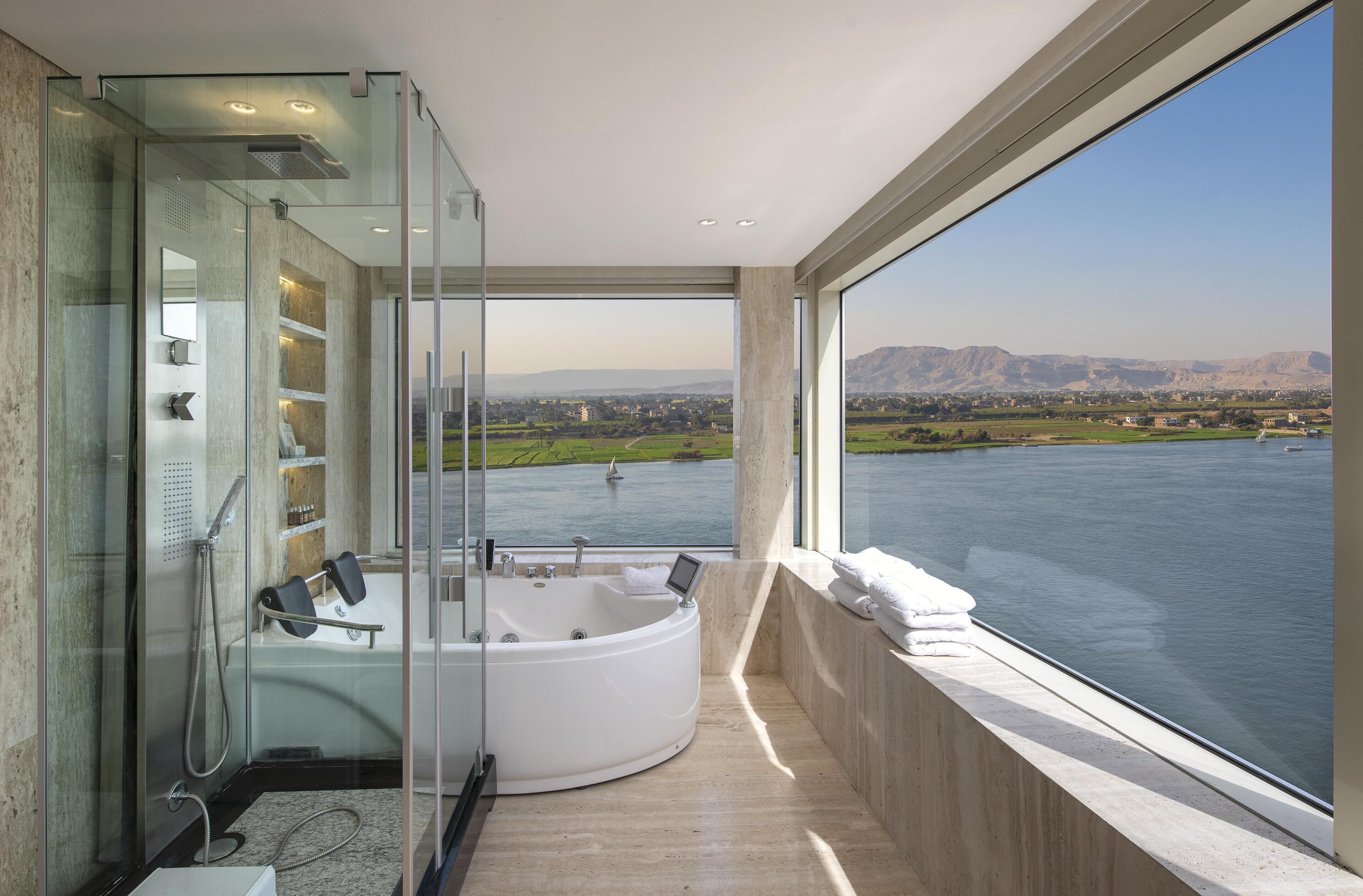 A modern bathroom with a glass-enclosed shower, a large bathtub, and a view of a body of water and distant landscape.
