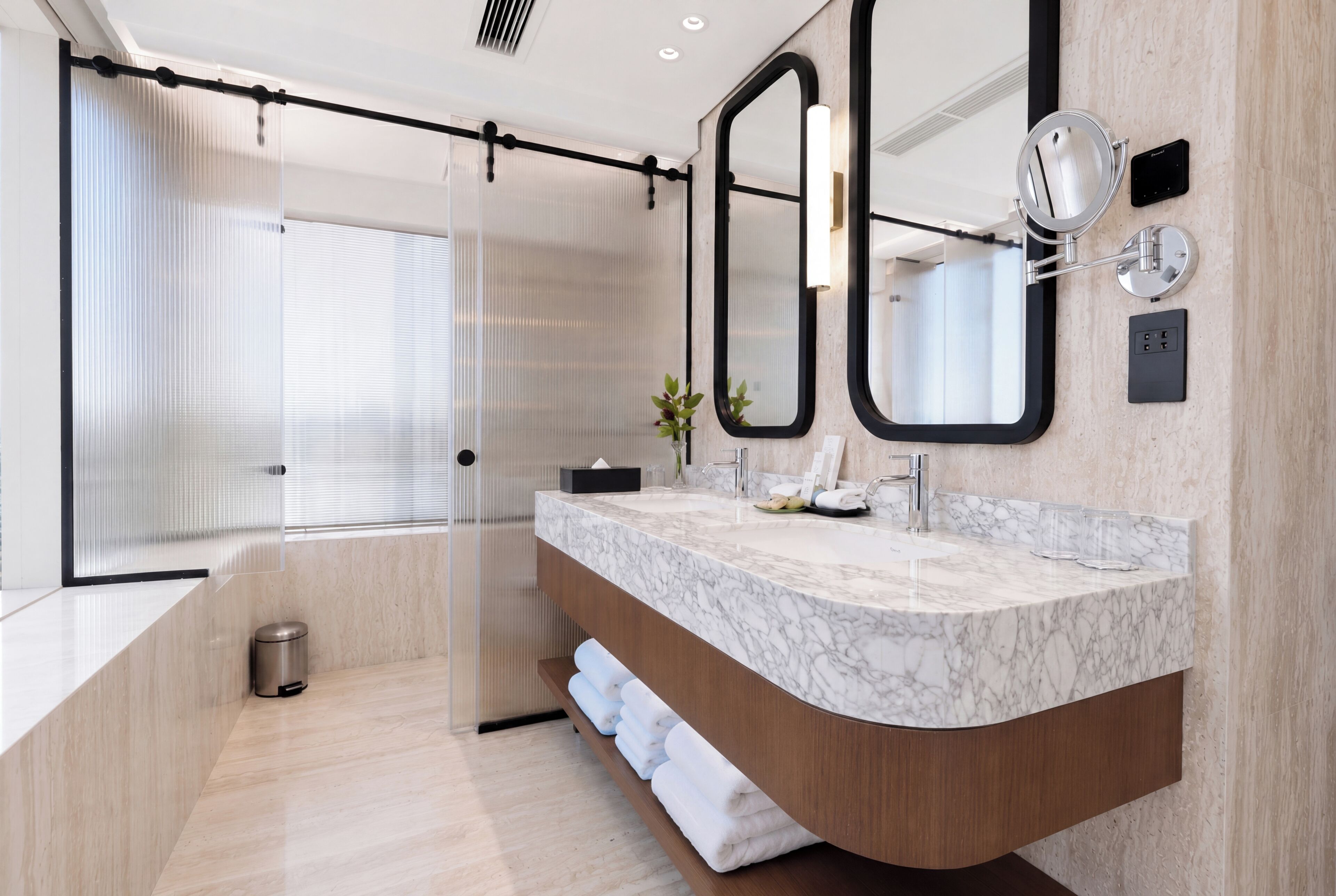 A modern bathroom with a marble vanity, a large mirror, a handheld mirror, and a glass shower enclosure.