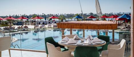 Un espace repas aménagé au bord de la piscine, avec des tables et des chaises, une vue sur la piscine et des parasols rouges et bleus.
