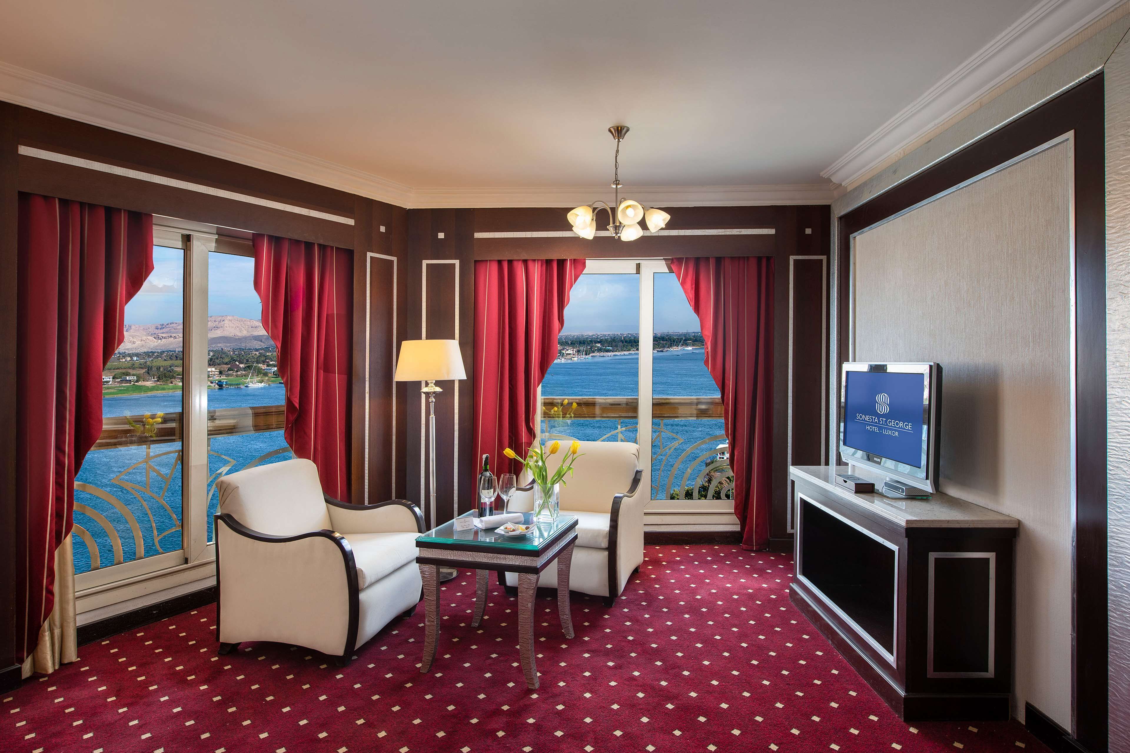 A hotel room with a large window, a television, a table with a vase of flowers, and a red carpet.
