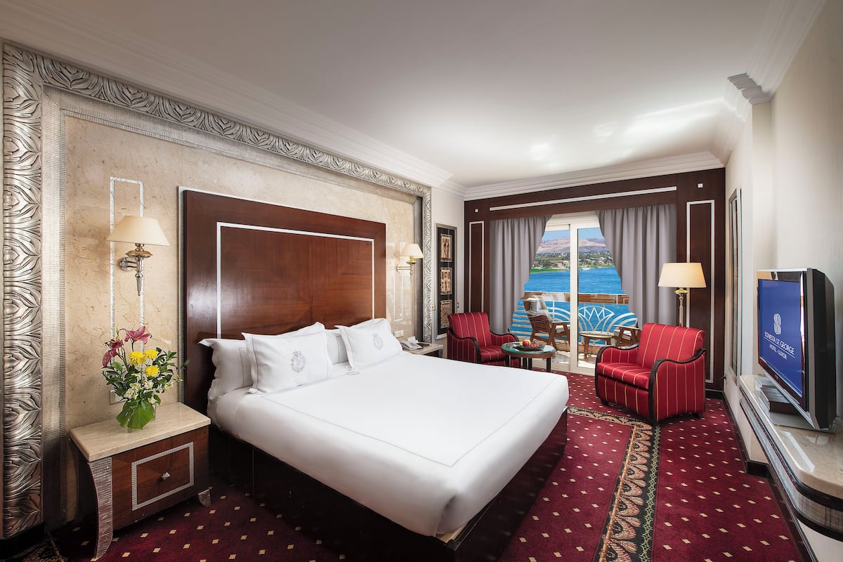 Quarto de hotel com uma cama grande, duas cadeiras, uma televisão e vista para o mar.