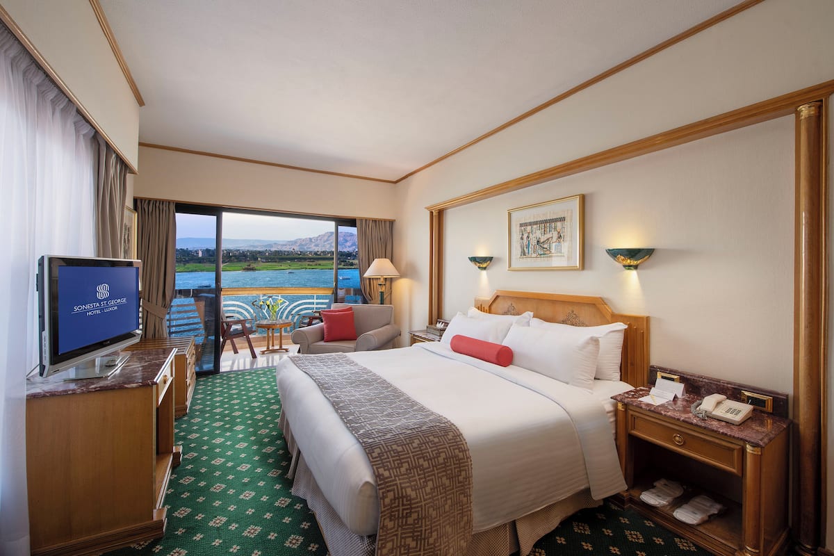 Quarto de hotel com cama, televisão, secretária com candeeiro, cadeira e vista para o exterior.