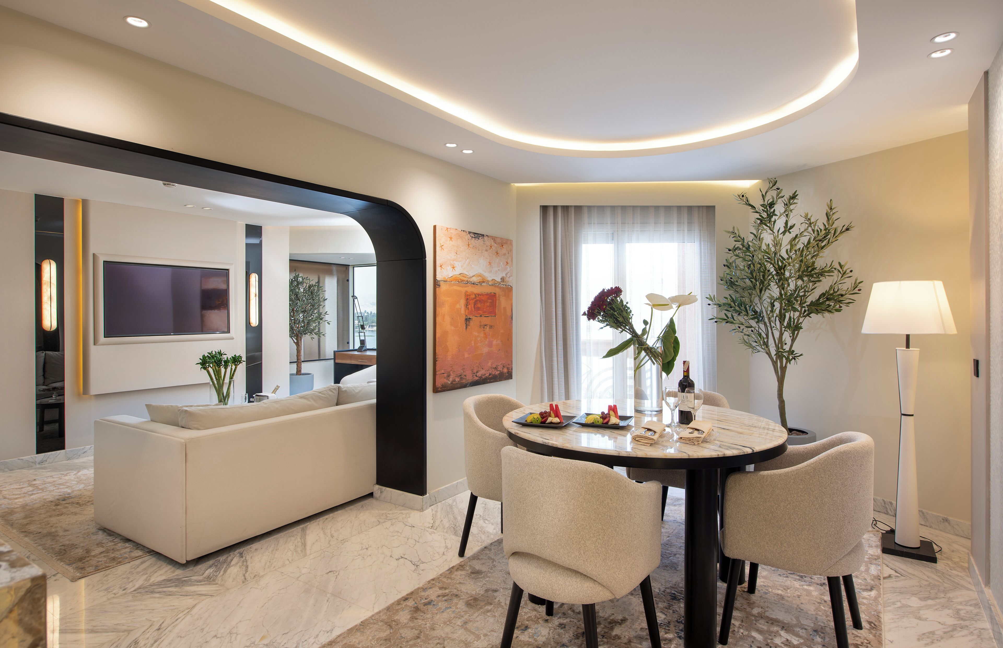 A modern hotel room with a dining area, a sofa, a flat-screen TV, and a large window with curtains.