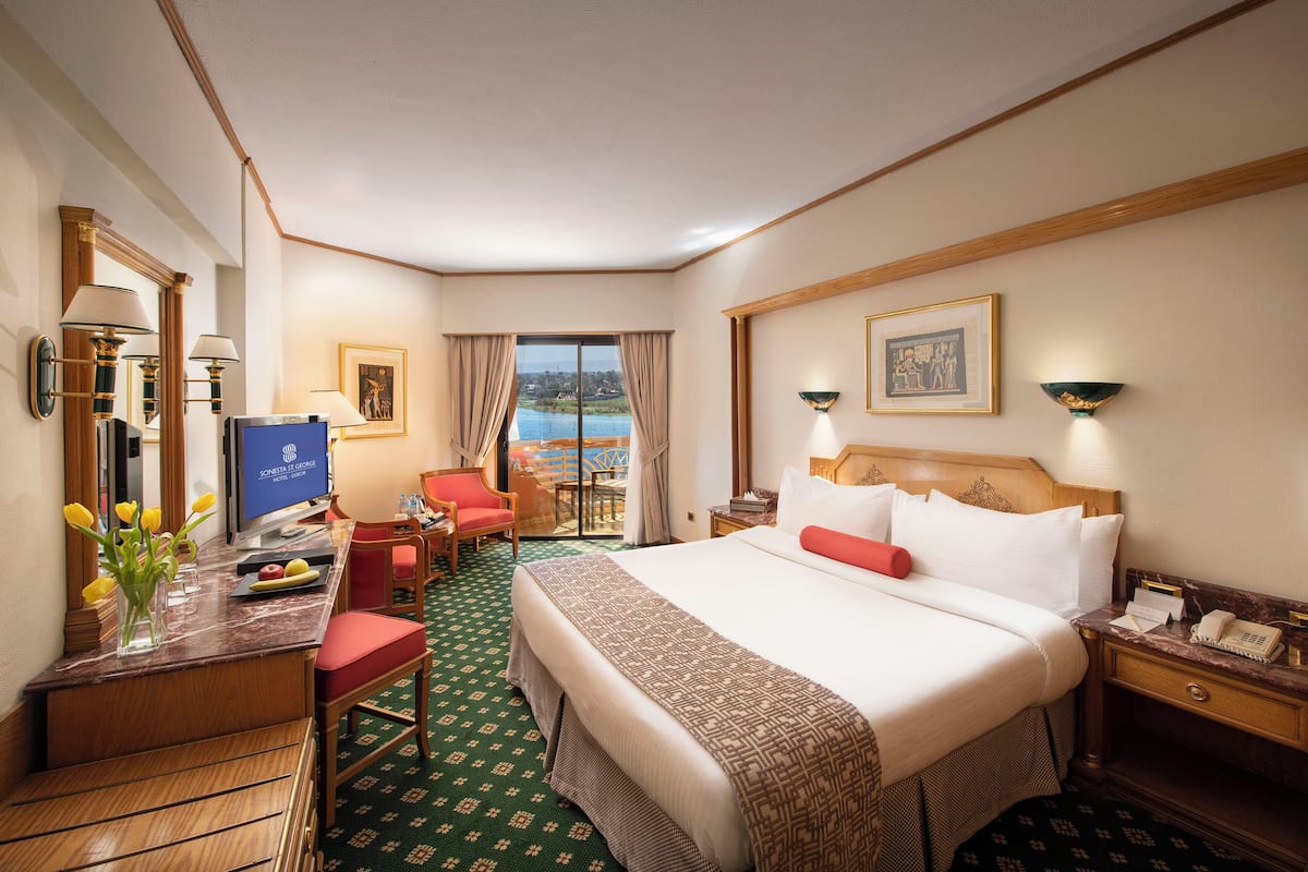 Quarto de hotel com uma cama grande, uma escrivaninha com computador, uma cadeira, uma mesinha com uma travessa de frutas e vista para o mar pela janela.