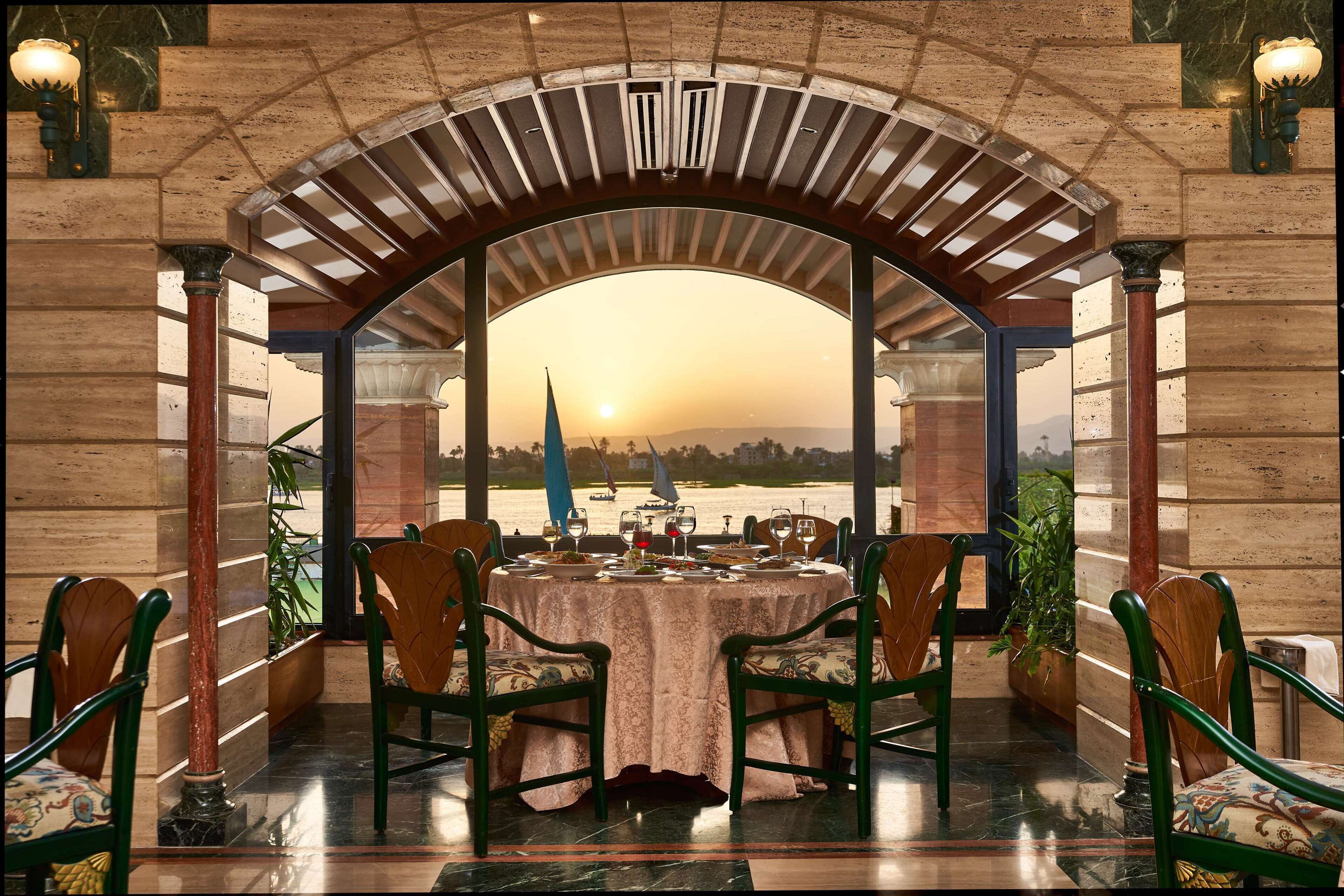 An outdoor dining area with a table set for four, surrounded by green upholstered chairs, under a stone archway with a view of the sunset over water.