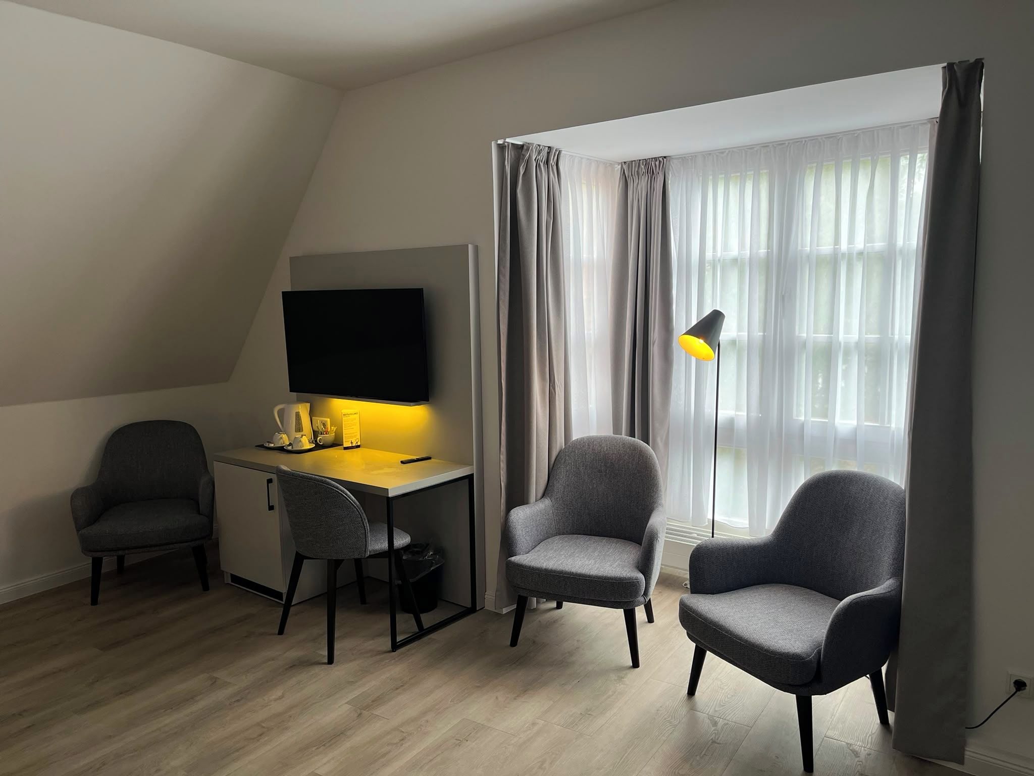 A modern room with a desk, two chairs, a flat-screen TV, a lamp, and a large window with curtains.