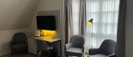 A modern room with a desk, two chairs, a flat-screen TV, a lamp, and a large window with curtains.