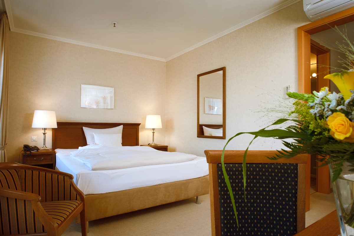 Une chambre d’hôtel avec un grand lit, deux tables de chevet avec des lampes, une chaise en bois, un miroir et un vase avec des fleurs.