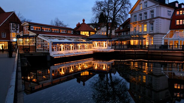 A riverfront building with a glass structure reflecting on the water, surrounded by other buildings. - DORMERO Hotel Bergström Lüneburg (Lueneburg)