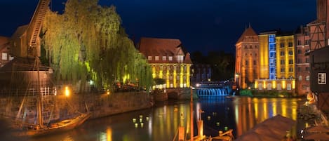 Blick auf eine historische Stadt am Flussufer, mit beleuchteten Gebäuden und einem traditionellen Boot, das an der Anlegestelle liegt.