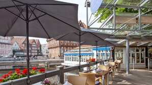 A wooden deck with tables and chairs, overlooking a canal with buildings in the background. - DORMERO Hotel Bergström Lüneburg (Lueneburg)