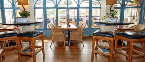 Un restaurante con mobiliario de mimbre, ventanas amplias y plantas en macetas.