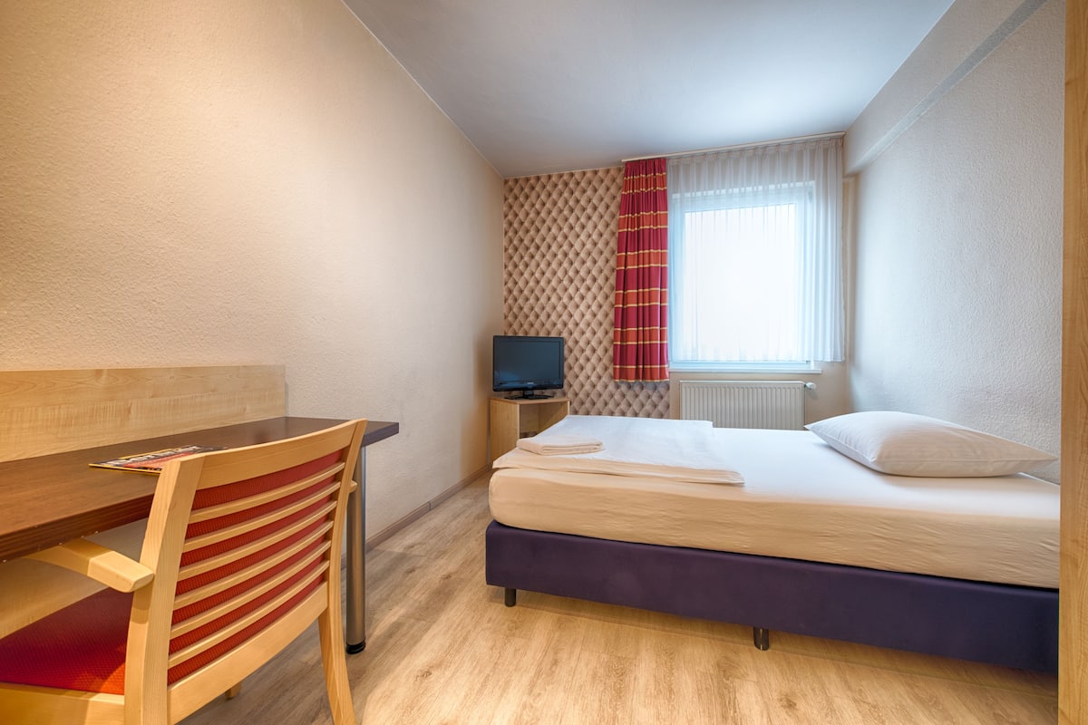 A hotel room with a bed, a desk, a chair, a television, and a window with curtains.