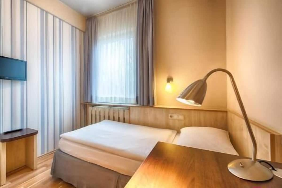 Et hotelværelse med seng, sengebord, lampe, fjernsyn og et vindue med gardiner.