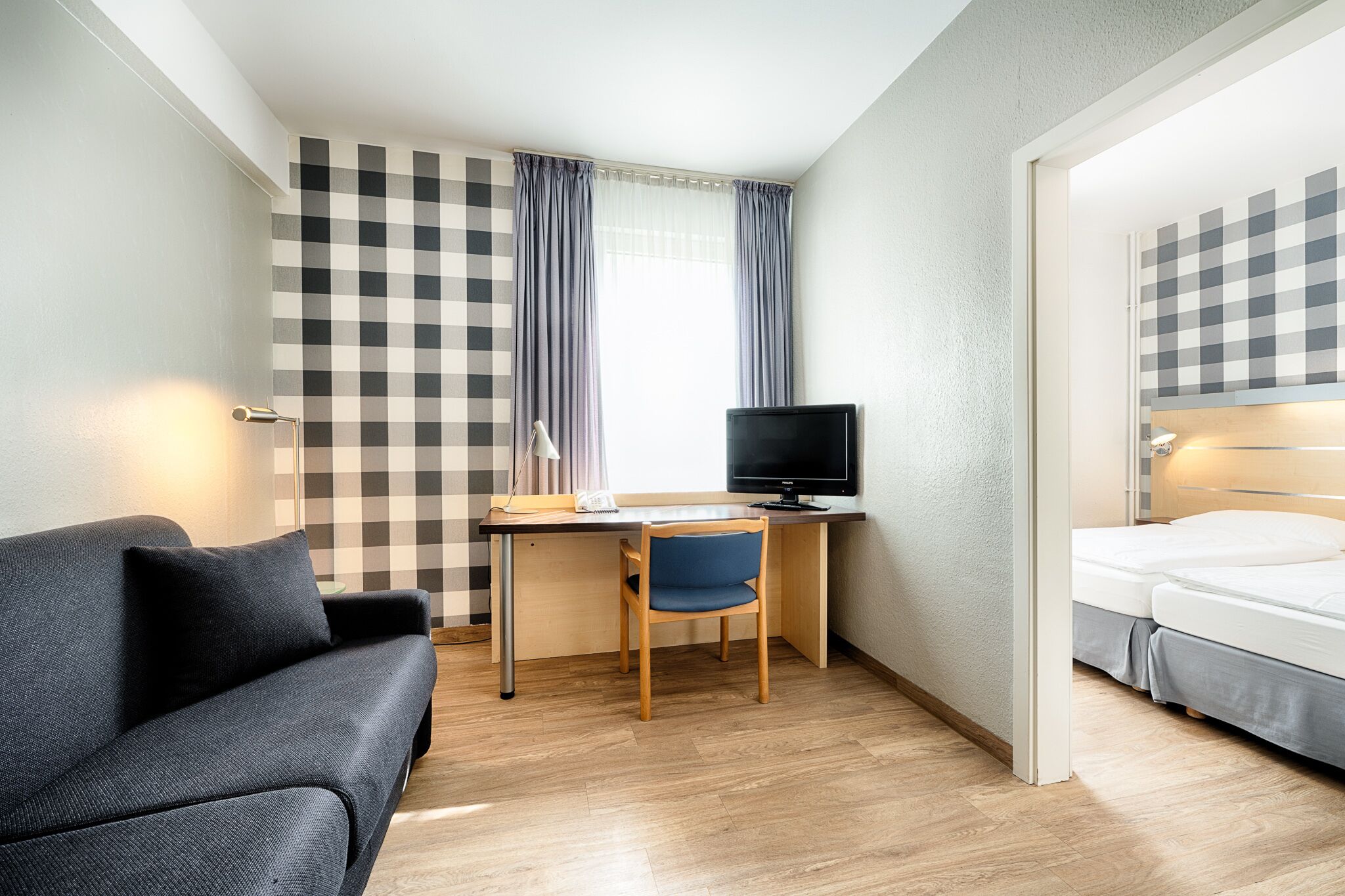 A hotel room with a checkered wall, a desk with a computer, a blue chair, a grey sofa, and a bed with a grey skirt.