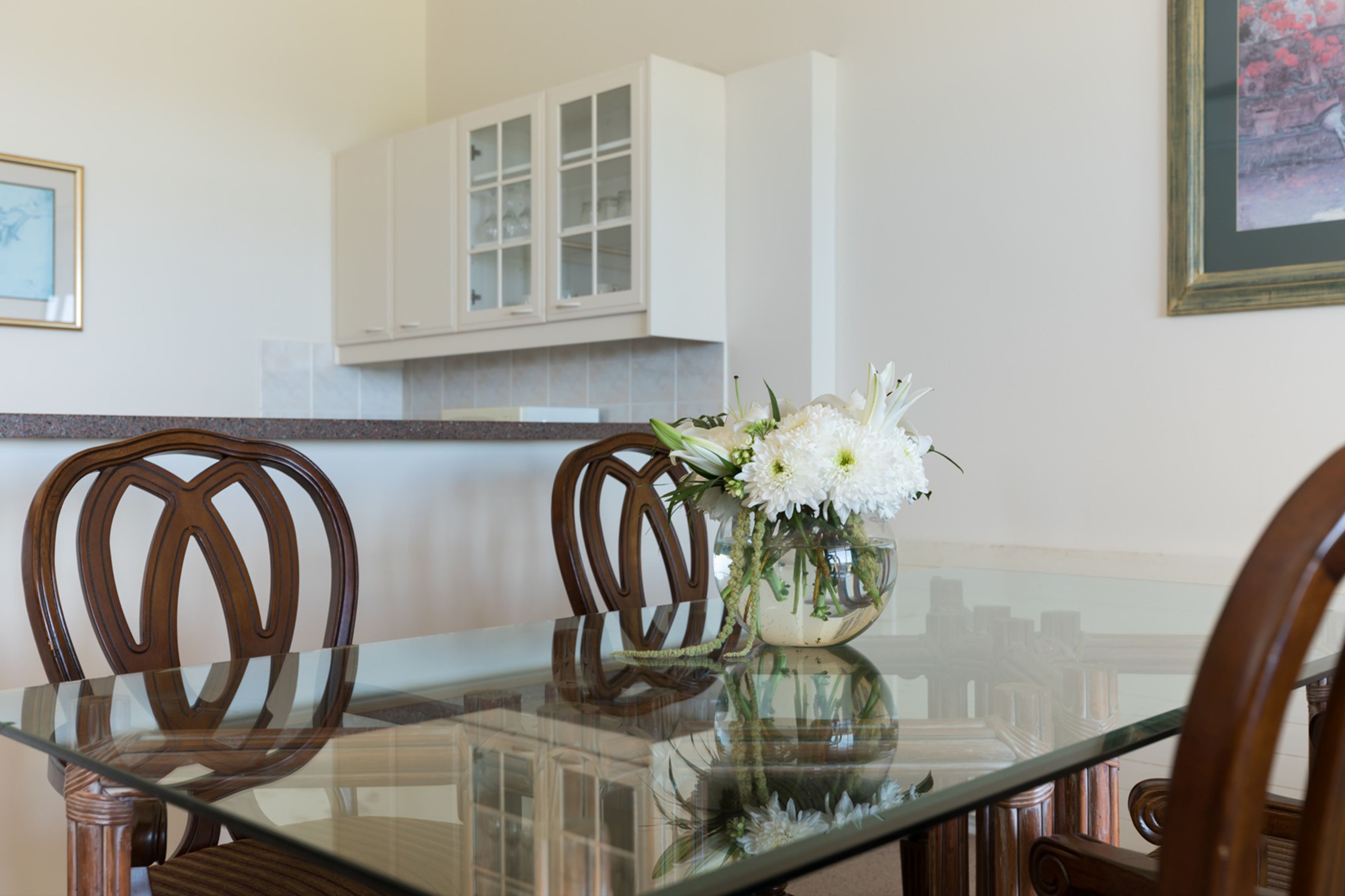 Un comedor con una mesa de vidrio, sillas de madera y un jarrón con flores blancas.