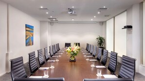 Un salón de conferencias con una larga mesa, sillas de cuero negro, un centro de mesa floral y un proyector en el techo.