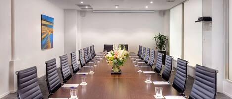 Un salón de conferencias con una larga mesa, sillas de cuero negro, un centro de mesa floral y un proyector en el techo.