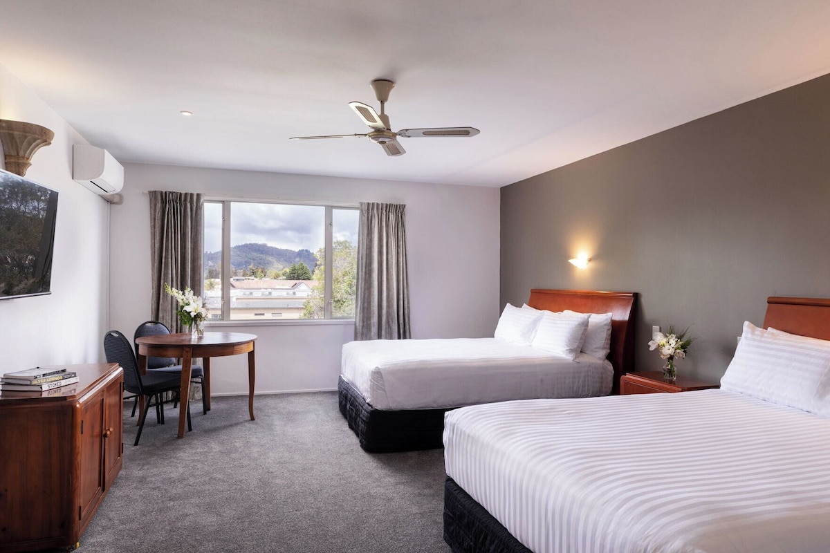 A hotel room with two beds, a desk, a chair, a ceiling fan, and a window with a view of the outdoors.