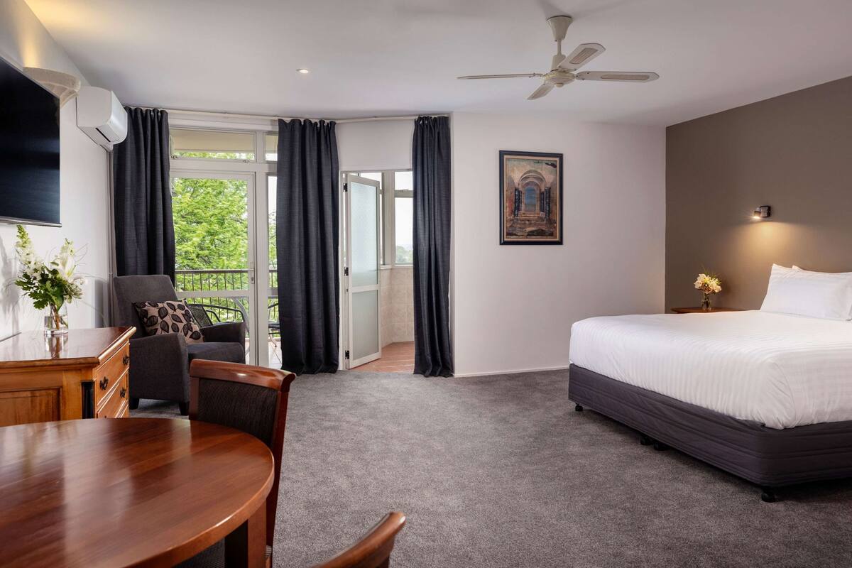A hotel room with a bed, a dining table, a chair, a ceiling fan, and a view of the outdoors.
