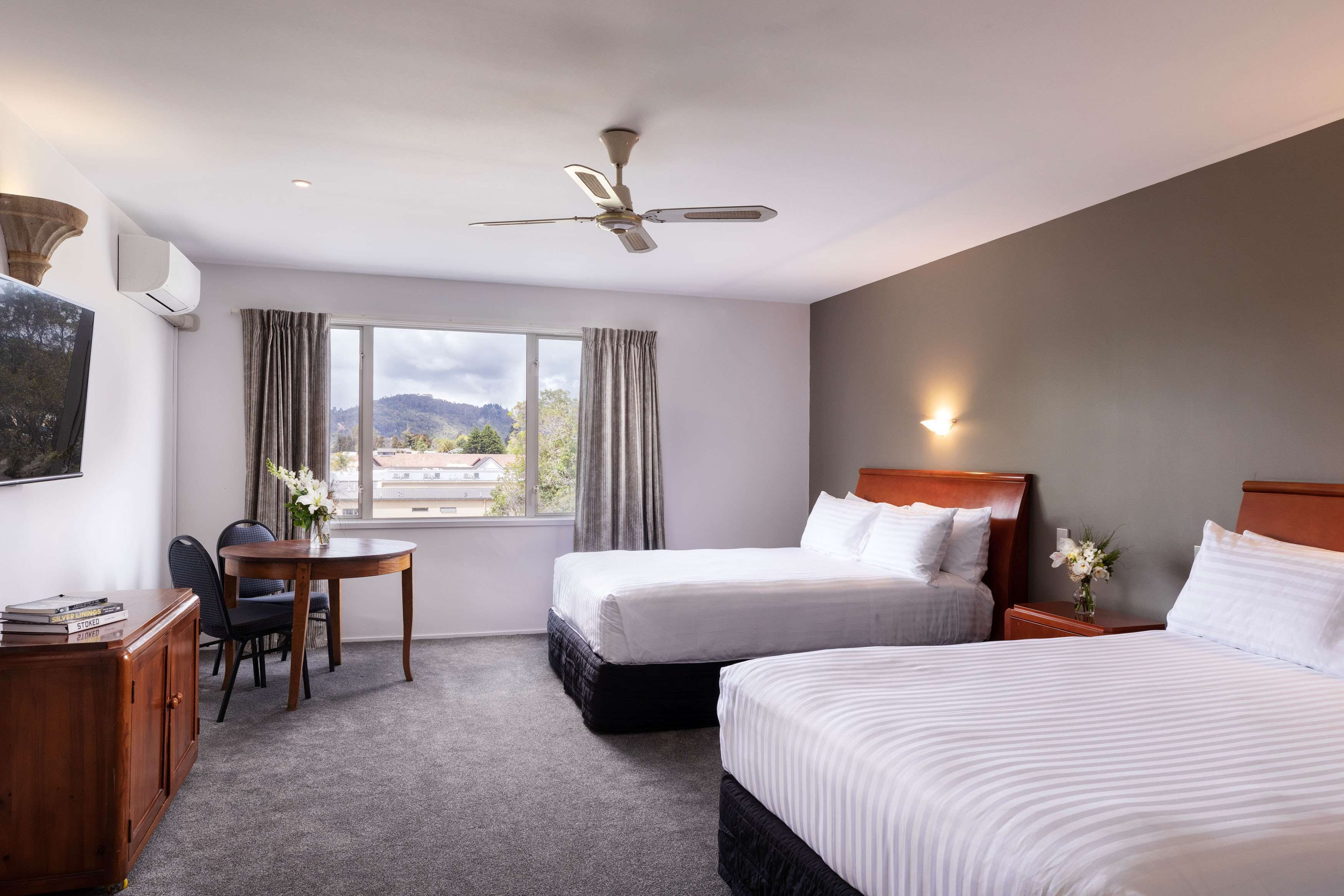 A hotel room with two beds, a desk, a chair, a ceiling fan, and a window with curtains.