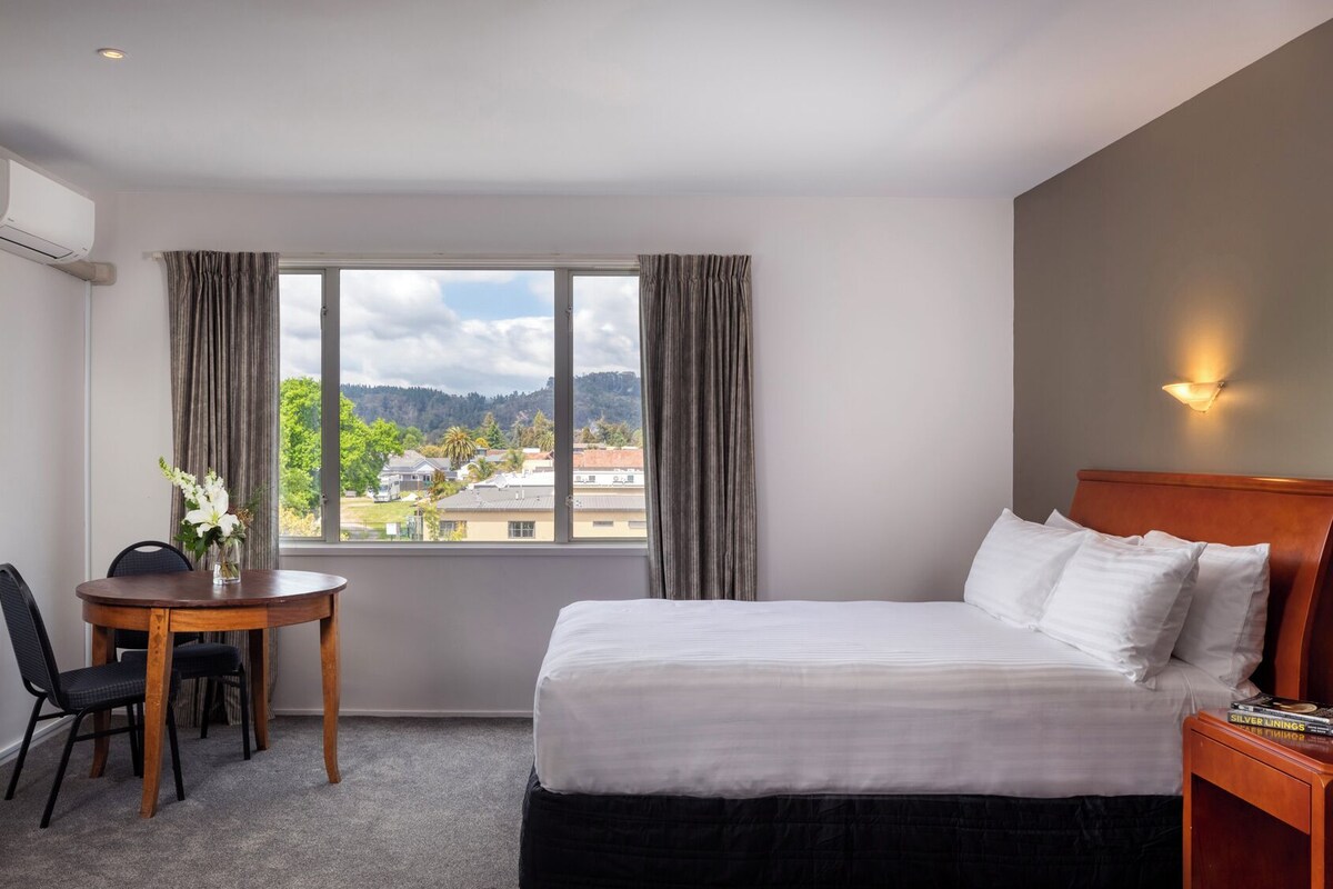 A hotel room with a bed, a desk with a chair, a window with curtains, and a view of a cityscape.